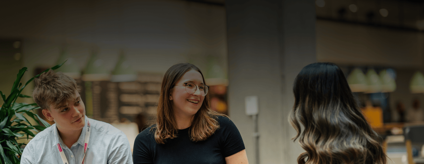 Three young adults talk inside a modern building; the woman in the centre smiles