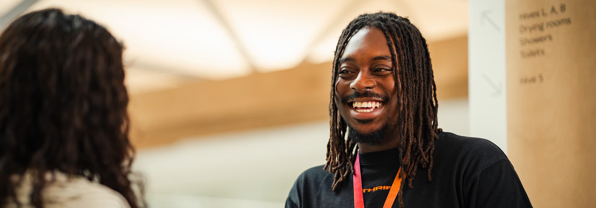 Man with dreadlocks smiling while talking to a woman in an office with warm lighting.
