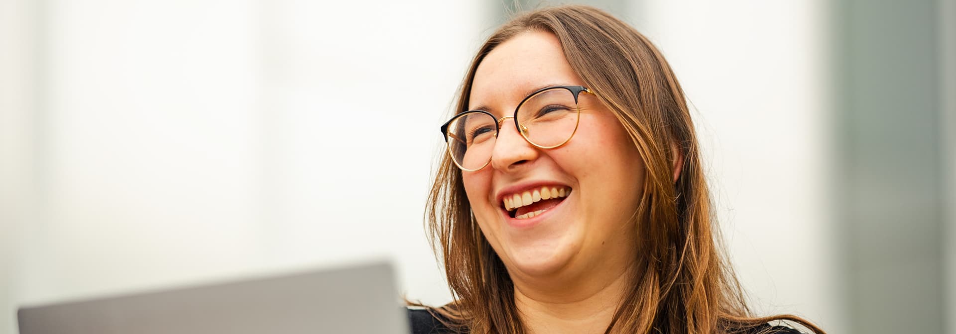 Woman with glasses laughing while working on a laptop outdoors