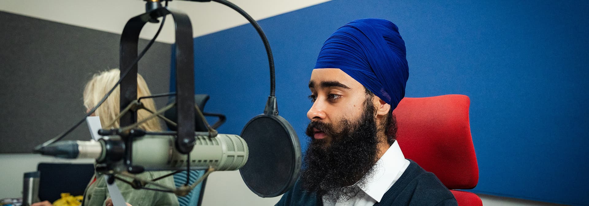 Man wearing a blue turban speaking into a microphone in a recording studio.