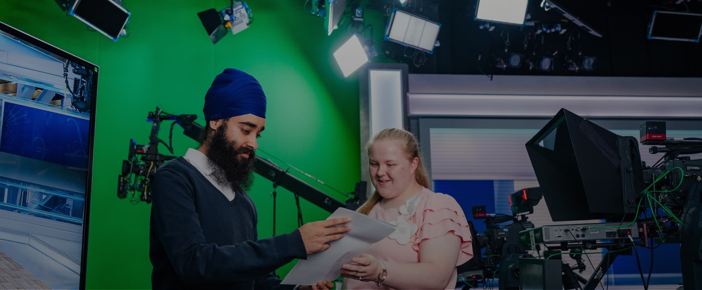 Two employees looking at paperwork in a television studio with green screen walls, ceiling rigging, and studio light.