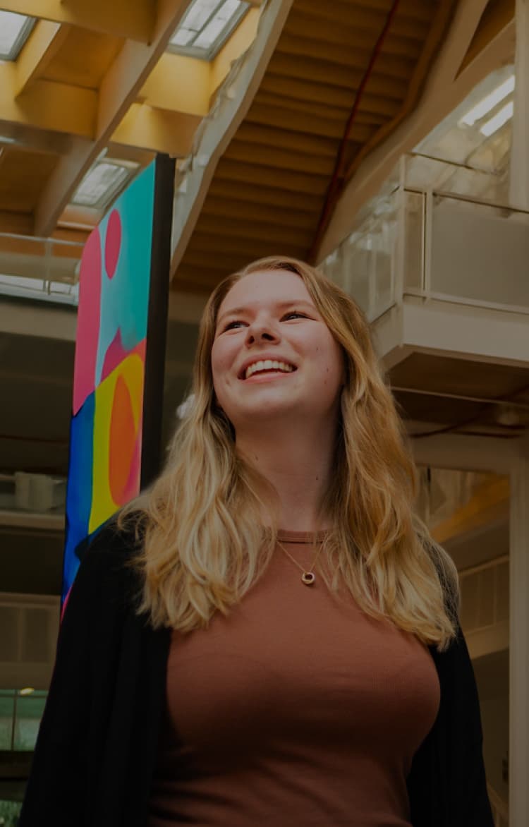Woman smiling and looking up in a brightly lit atrium with colorful abstract artwork behind her.