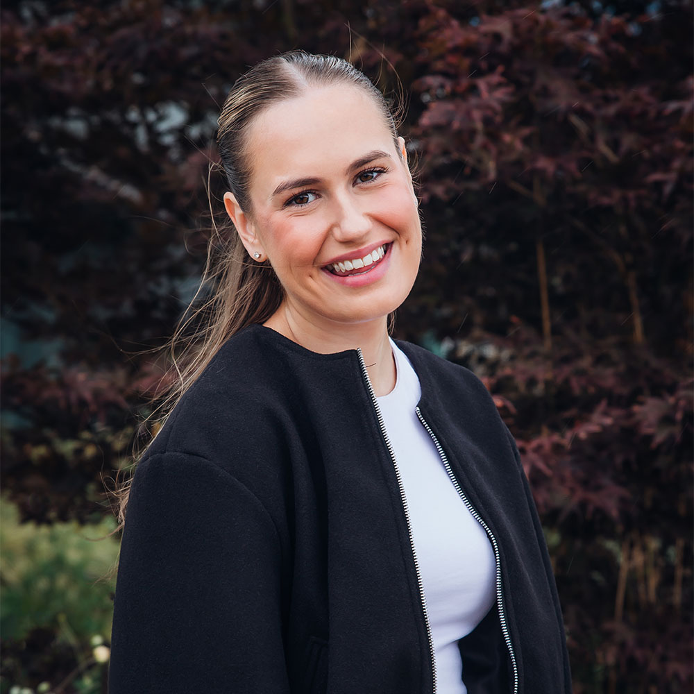 Tilly, wearing a black jacket and a white t-shirt smiling, with green and dark red leaved trees in the background