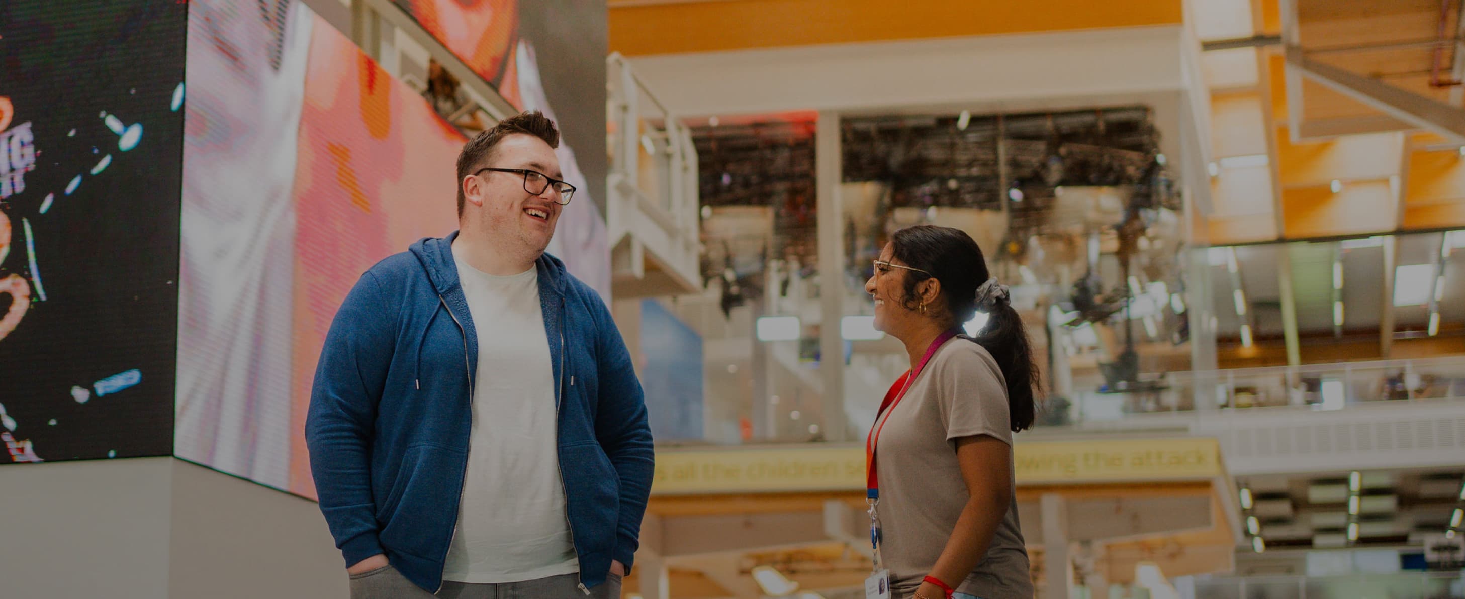 A man in a blue hoodie and woman with a red lanyard smiling and talking in an open, tech-style atrium.