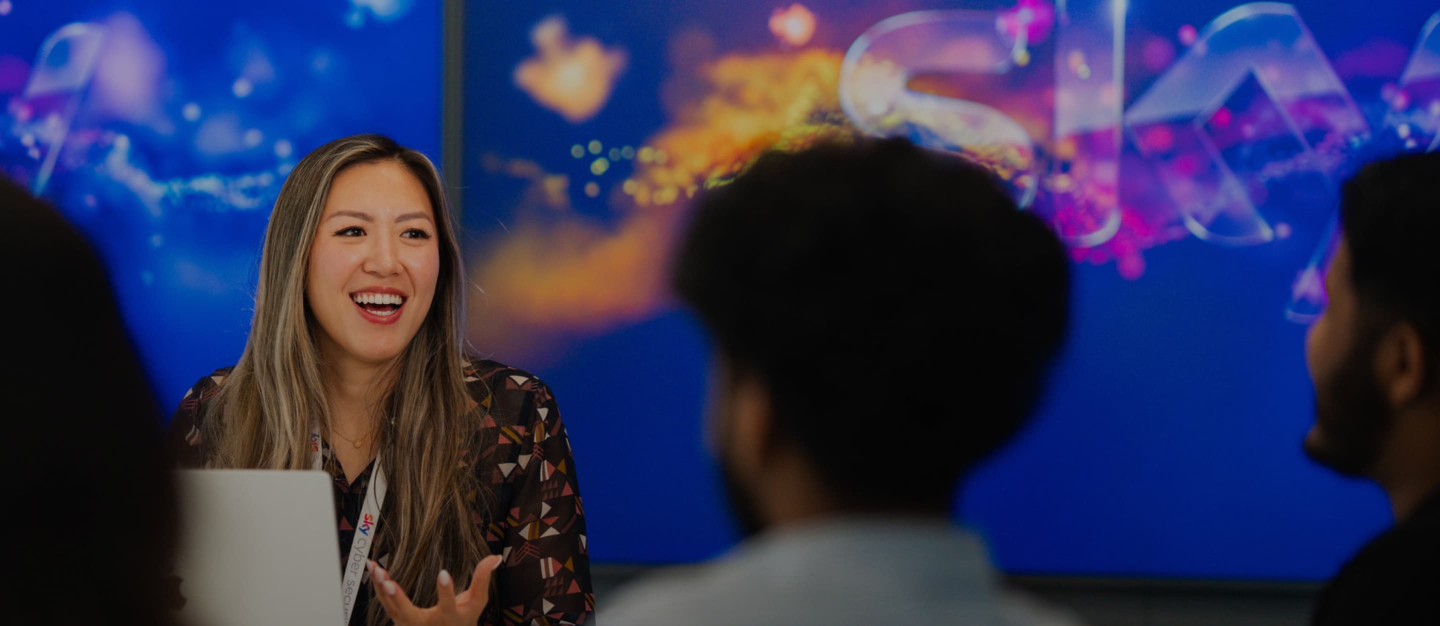 Woman with long hair and patterned shirt smiling and gesturing while seated at a laptop in front of a blue display.