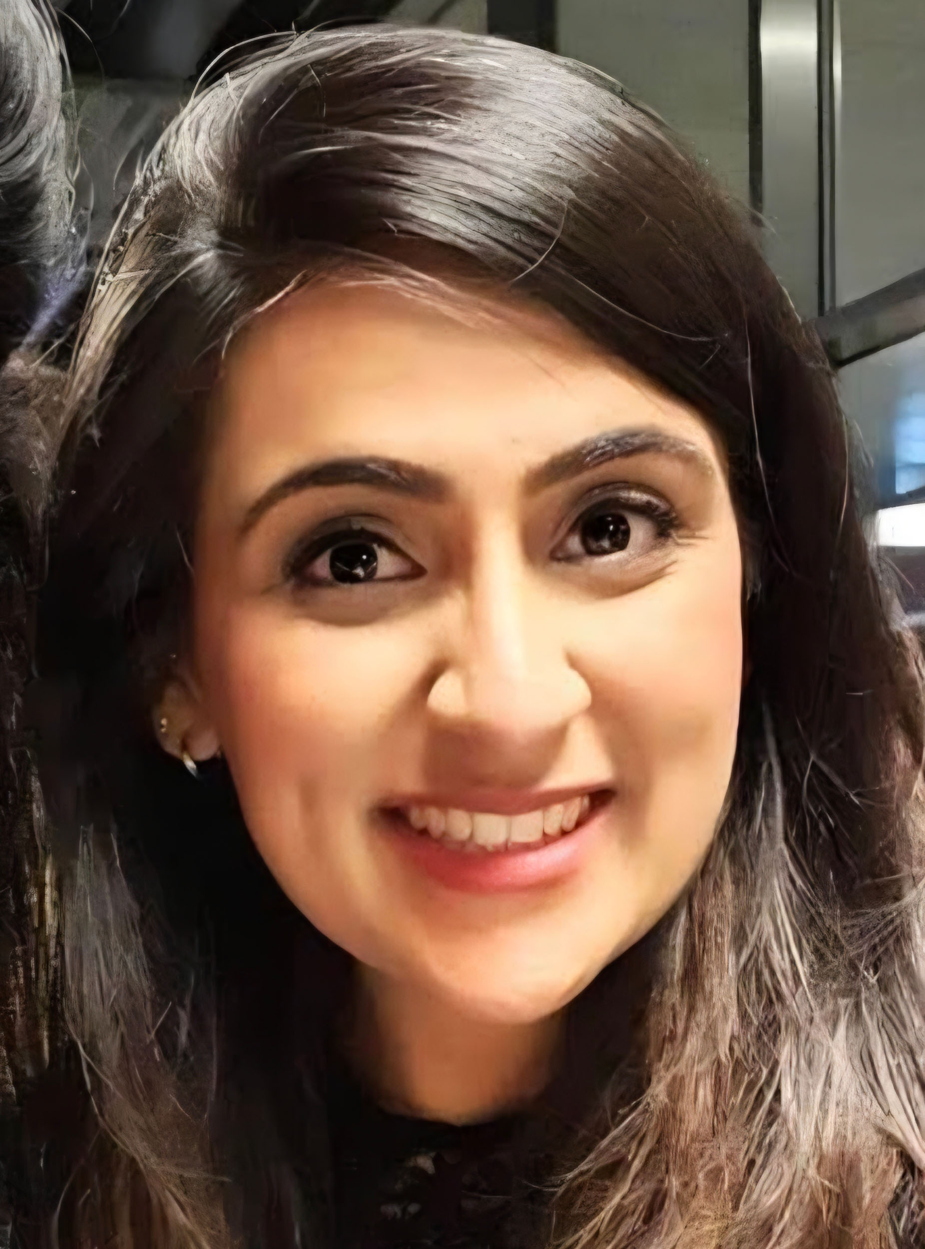 A close selfie shot of Meera looking into the camera lens. Meera is smiling - next to the frame of a window. Meera has shoulder length dark hair.