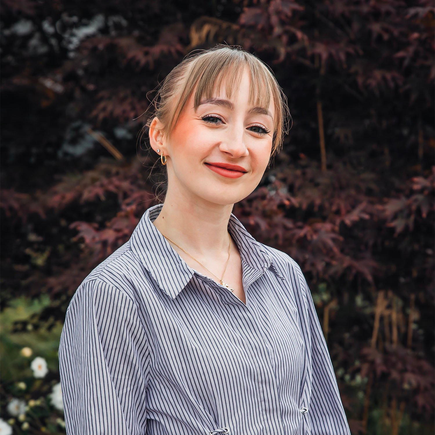 Chloe, wearing and blue and white striped shirt, smiles as she looks towards the camera. Behind Chloe is a vibrant scene of green and dark red leaves.