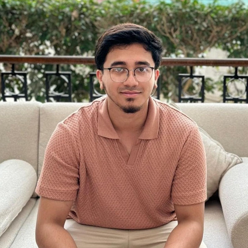 Syed, looking into the camera, sits forward on a cream sofa. He's wearing a salmon short-sleeve shirt, and beige trousers. Behind Syed is a row of bright green bushes.