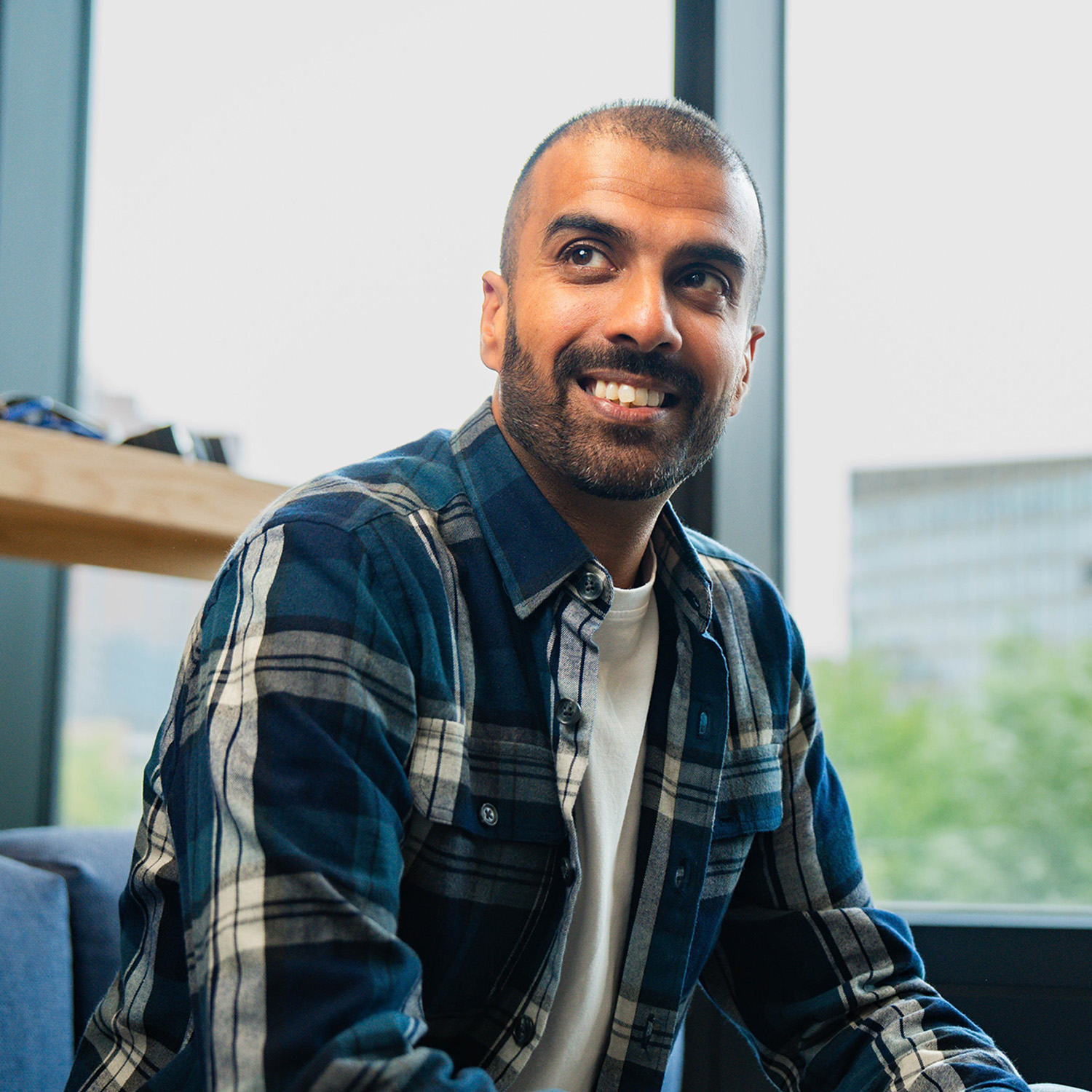 Parmjit smiles and looks off to the left of the photo. He wears a blue and white checked button shirt with a white t-shirt, and sits on a blue sofa. In the background the edge of a wooden deskspace, in front of a large panel window overlooking a large office block and trees in the distance.