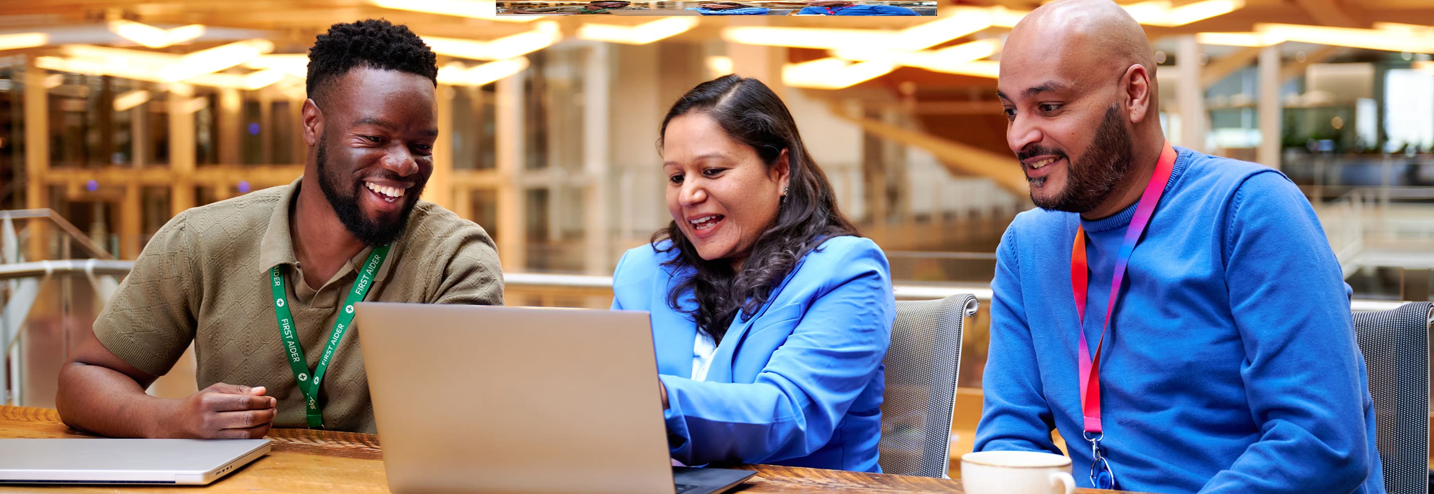 Three colleagues smiling and discussing something on a laptop in a bright office setting.