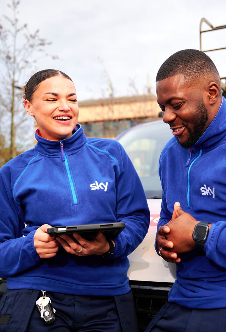 Two Sky engineers in blue uniforms smiling and reviewing something on a tablet outdoors.