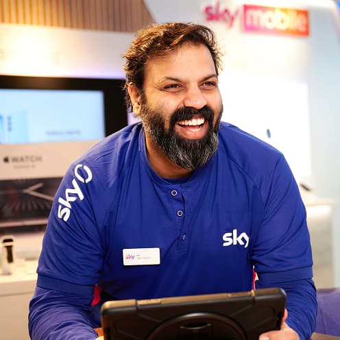 Sky retail staff member in uniform smiling behind a counter with tech products in the background.