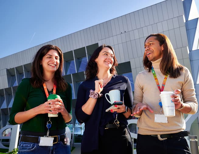 Three colleagues laughing and holding drinks outside a modern glass building on a sunny day.