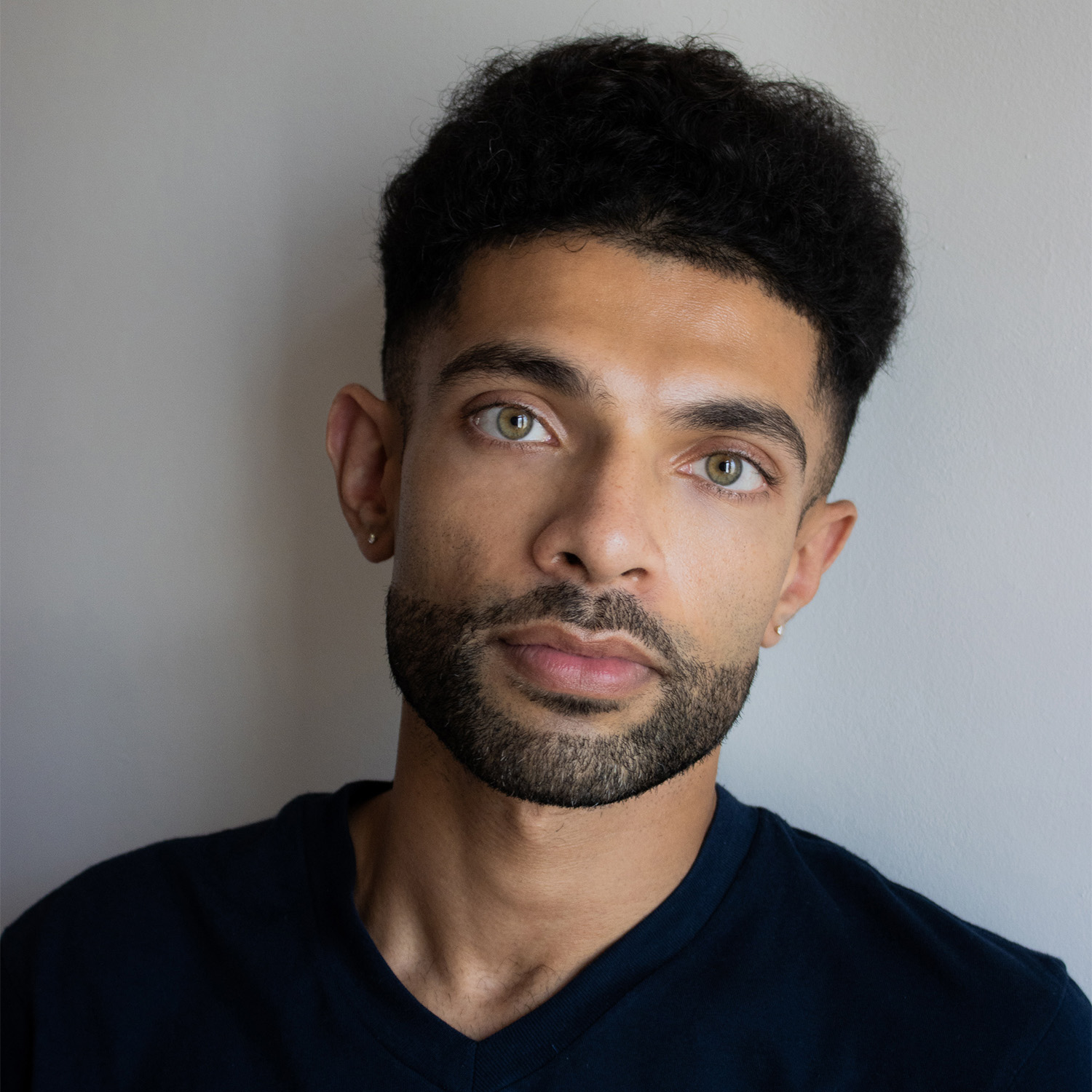 Joyston looks directly into the camera, with his head tilted slightly to the left. He is wearing earrings in both ear lobes, and a black v-neck t-short. He is standing in front of a plain white wall, with his shadow just behind him.