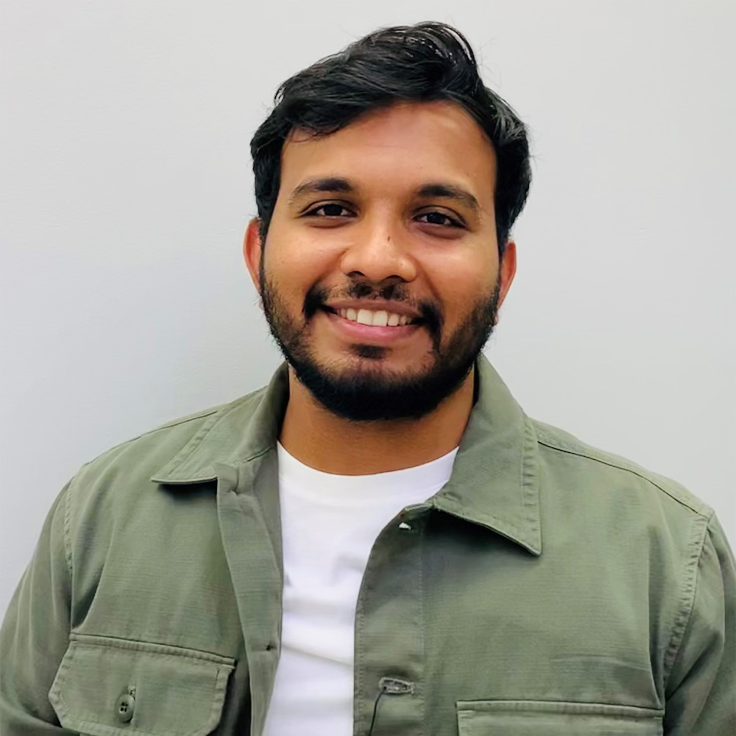Vishnu looks into the camera with a smile. He is stood in front of a white wall, and is wearing a khaki-green overshirt and a white t-shirt.