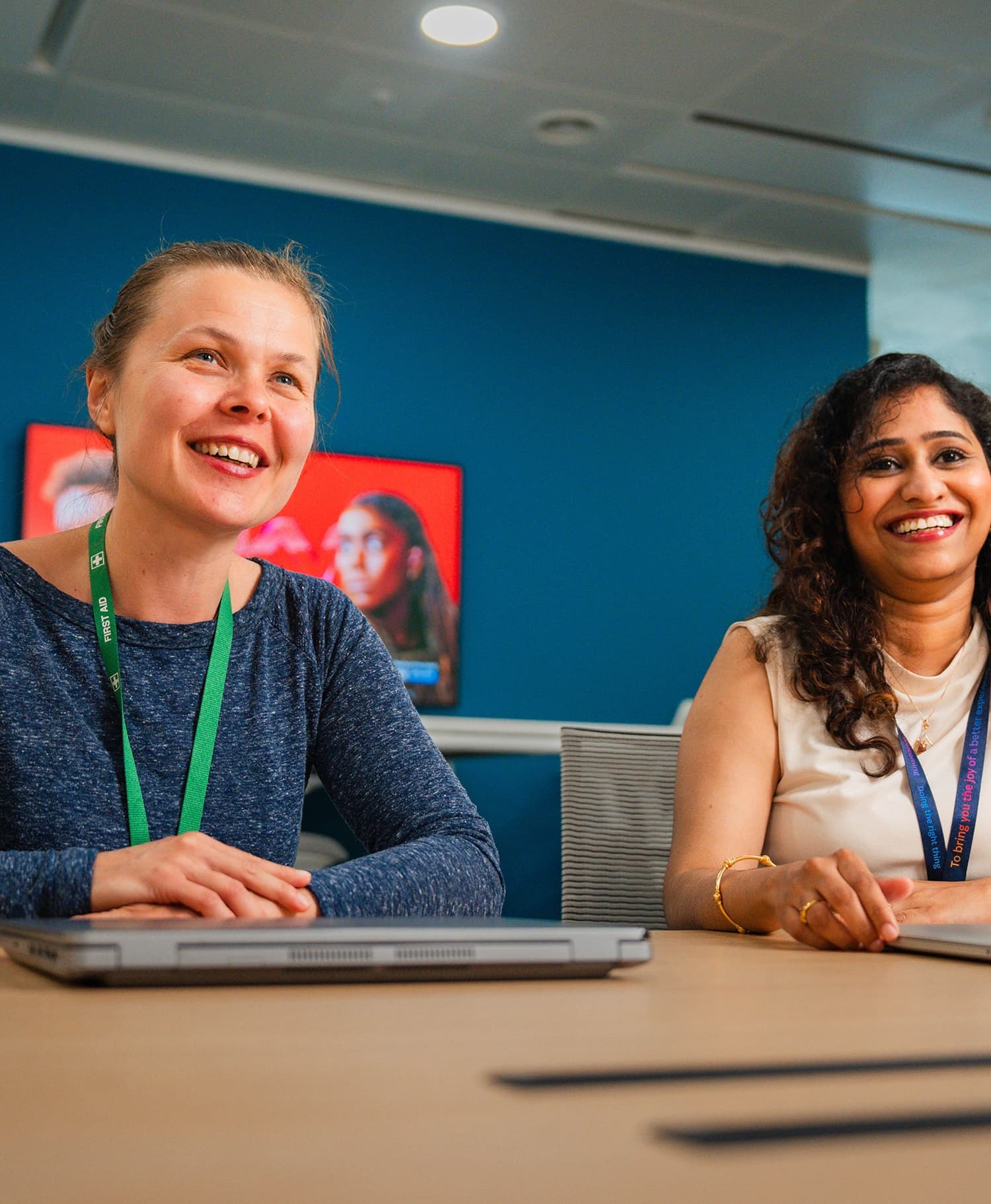 Two women seated at a conference table with laptops, smiling in a modern meeting room.