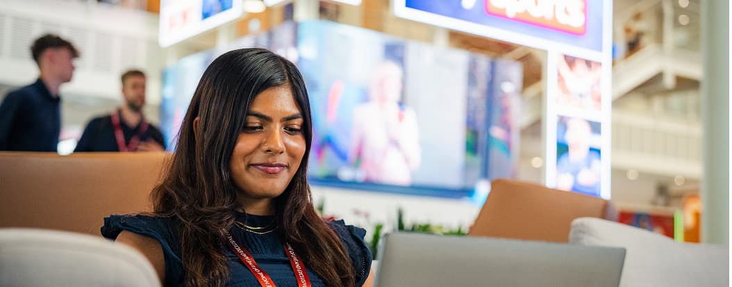 Woman wearing a Sky lanyard works on a laptop in a modern office with screens and lights in the background.