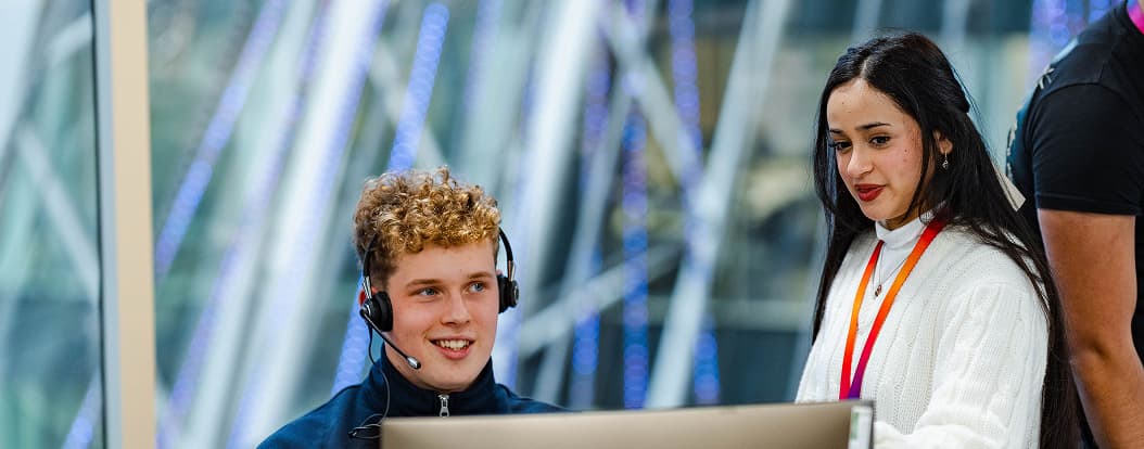 Two colleagues collaborate at a computer; man wearing a headset sits while woman with lanyard stands beside him.