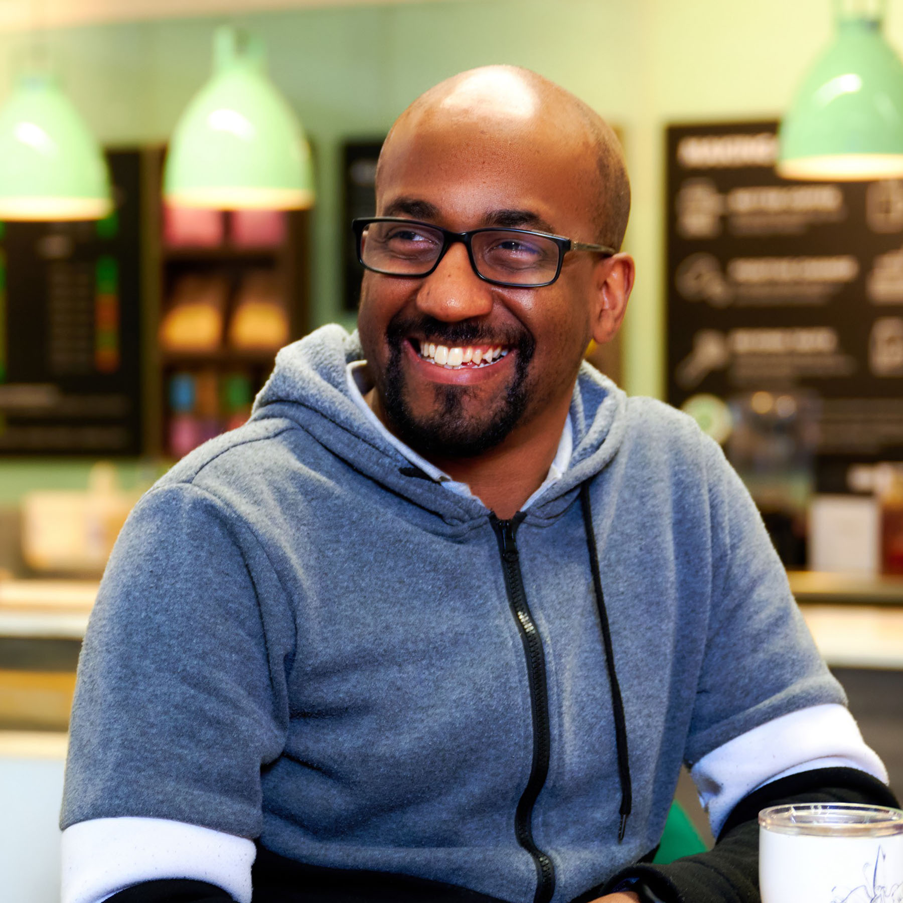 Nathan is sitting indoors at a café-style table. Nathan looks to the left of the camera, wearing a soft, heather blue zip up hoodie with dark drawstrings. In front Nathan, on the right side of the frame, there is a Sky branded coffee mug. The background shows a brightly lit counter area with mint green hanging pendant lights, menu boards with chalkboard-style text, and blurred café equipment. The overall setting looks casual and warm, with soft lighting and muted colours.