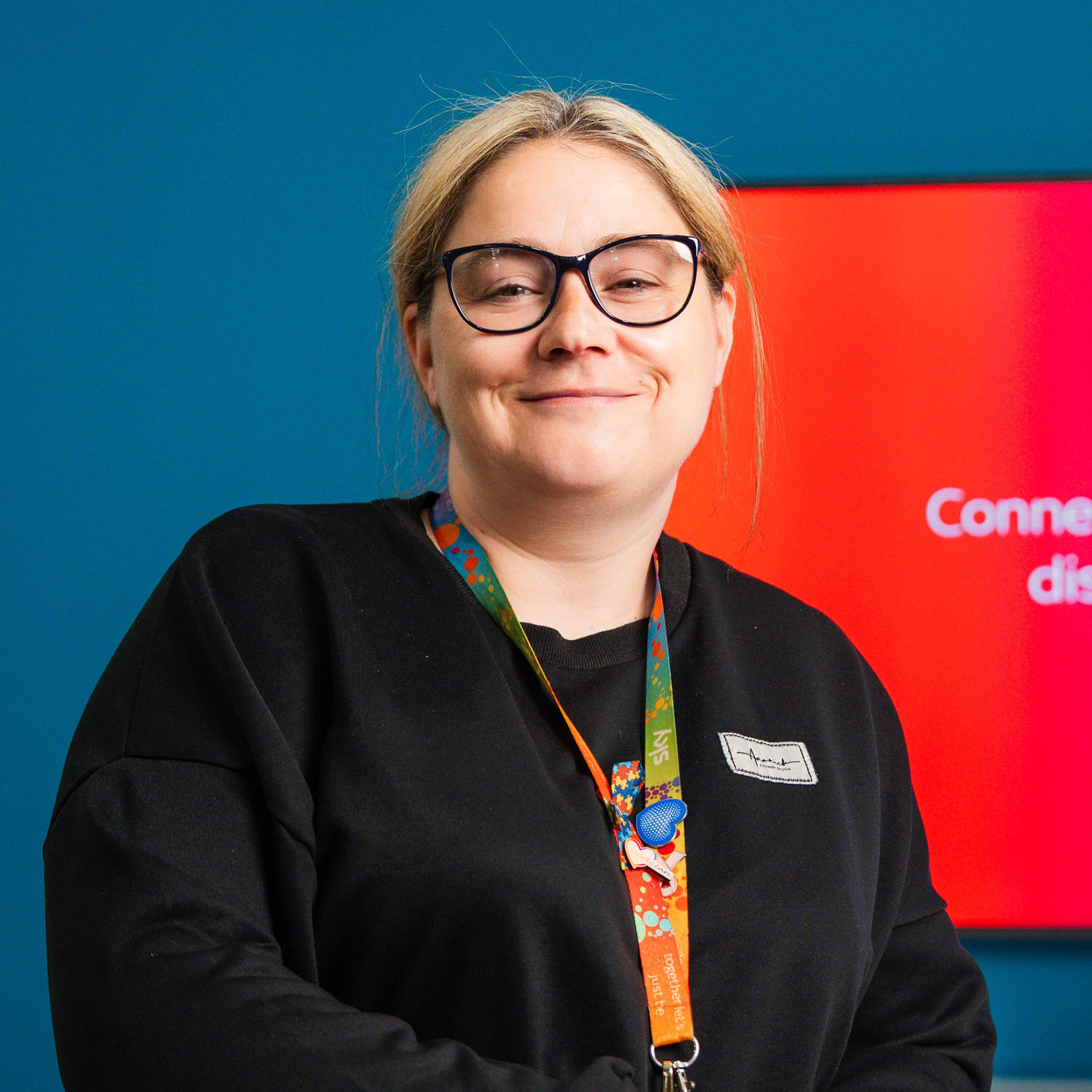 Nicola stands indoors beside a white desk and office chair, against a teal wall. Nicola wears a pair of glasses, a black long sleeve top and a patterned lanyard. Behind them, a wall mounted screen shows a red to pink gradient with the text “Connect your HDMI device to display your content.”
