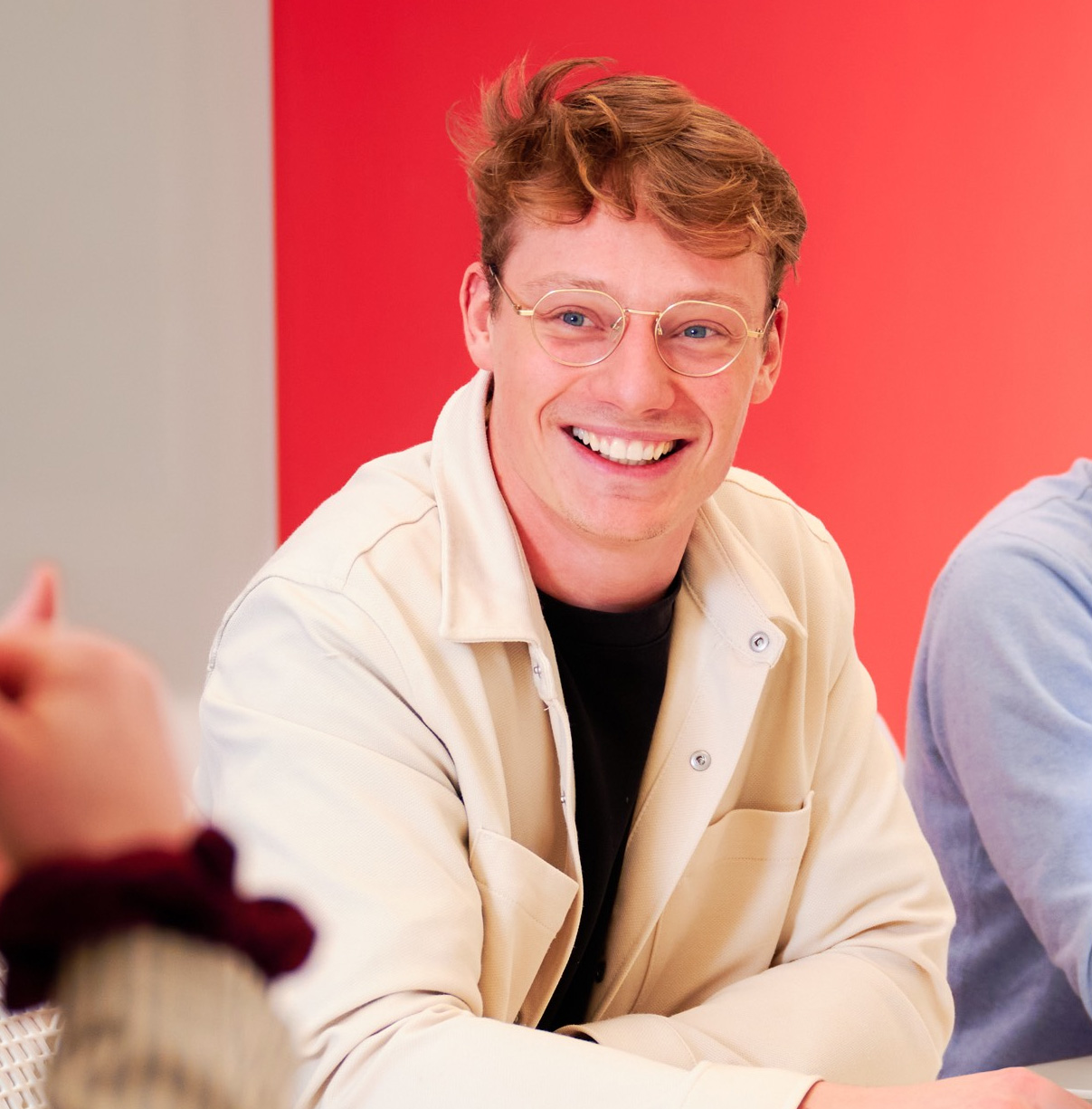 Fraser is seated indoors at a table, wearing a light beige overshirt layered over a dark top. Another person’s hand and sleeve are visible in the foreground on the left side. The background features a wall split between light grey on the left and bright red on the right. The scene has soft, even lighting and takes place in a collaborative meeting.