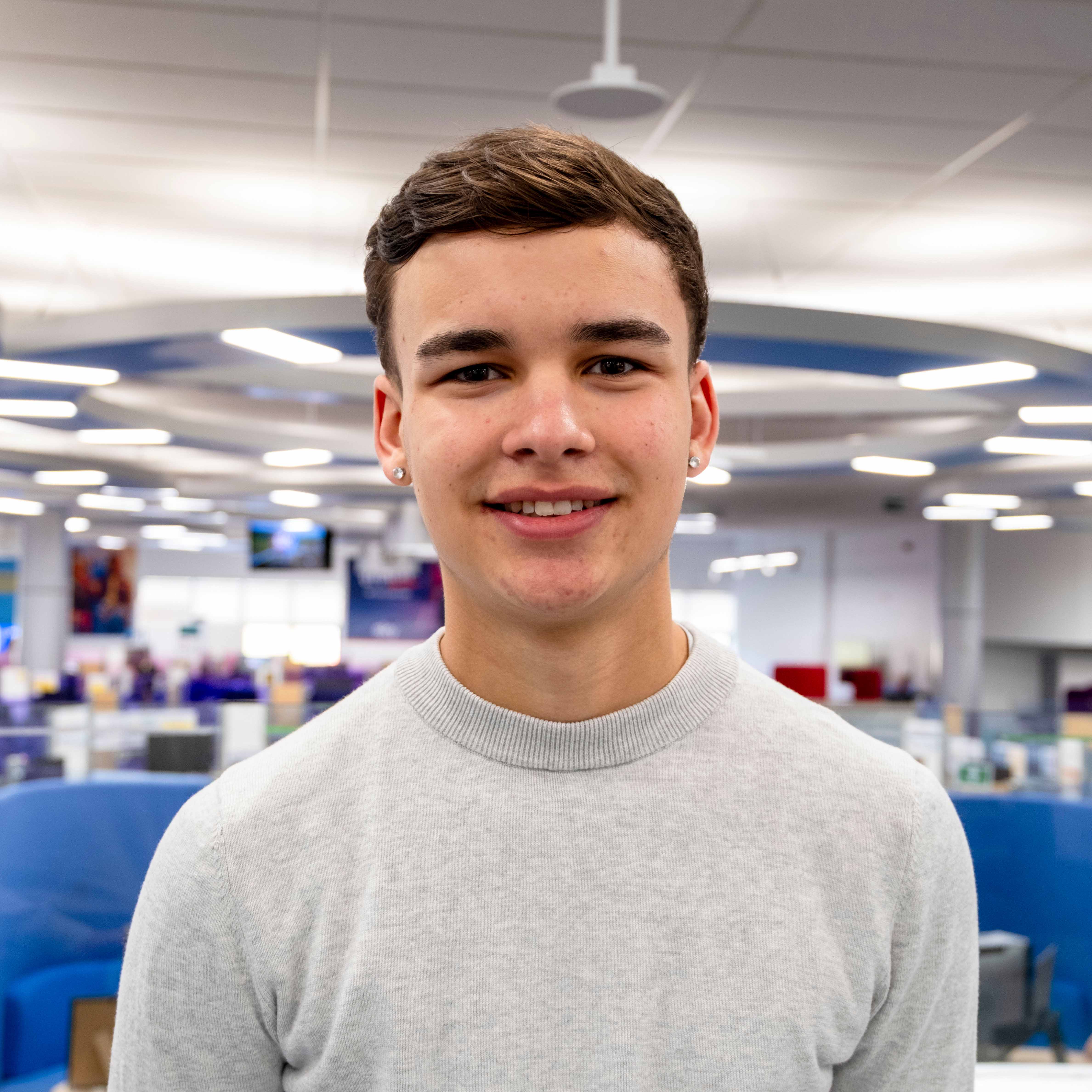 Isaac, wearing a grey crew neck jumper and a set of pear earrings, smiles as he looks towards the camera. Behind Isaac is a vibrant scene of the office.