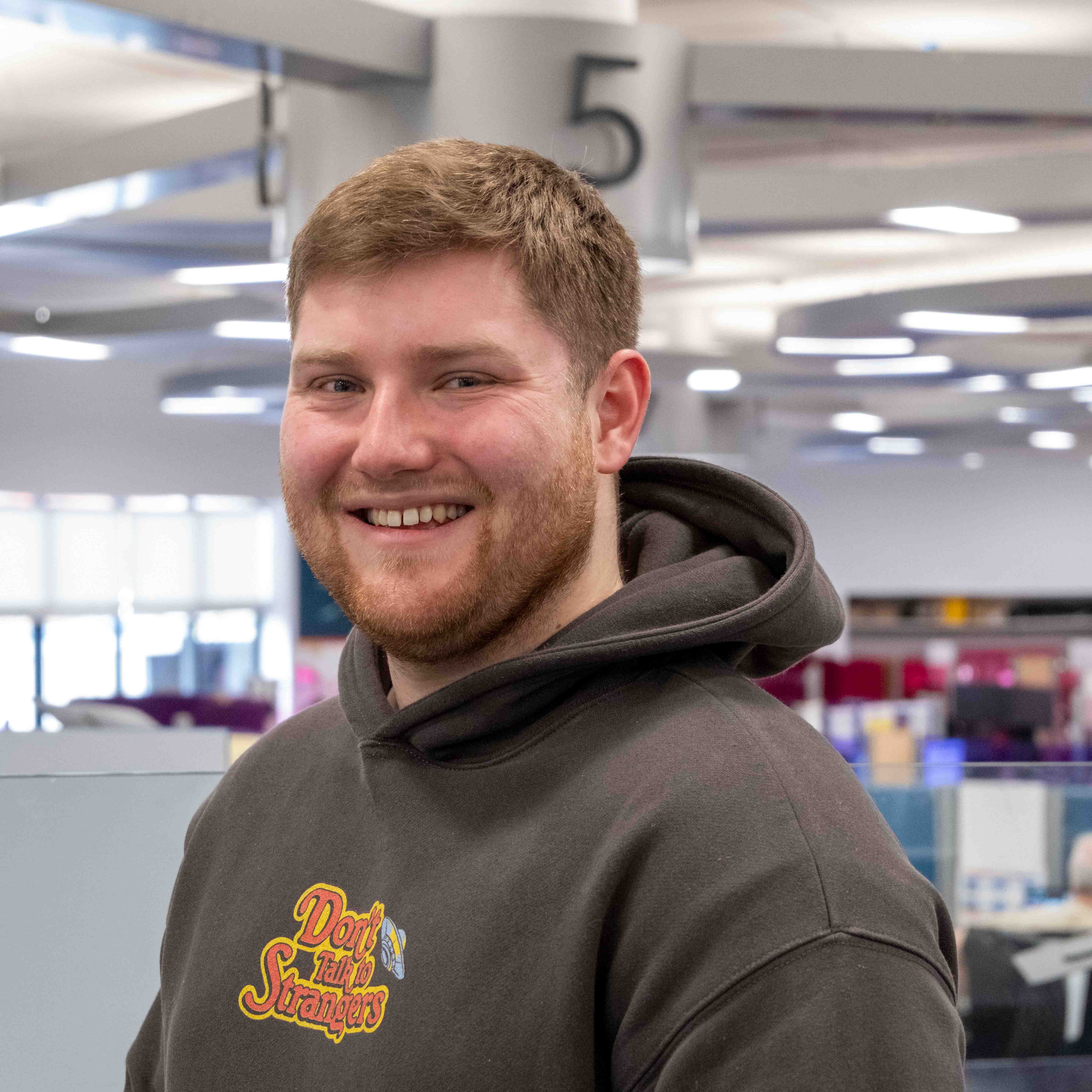 Jake, Wearing a dark beige hoody with an orange logo in the middle. Huge smile on his face, housy brown hair with vibrant office behing him and a big 5 to show which department area they are in