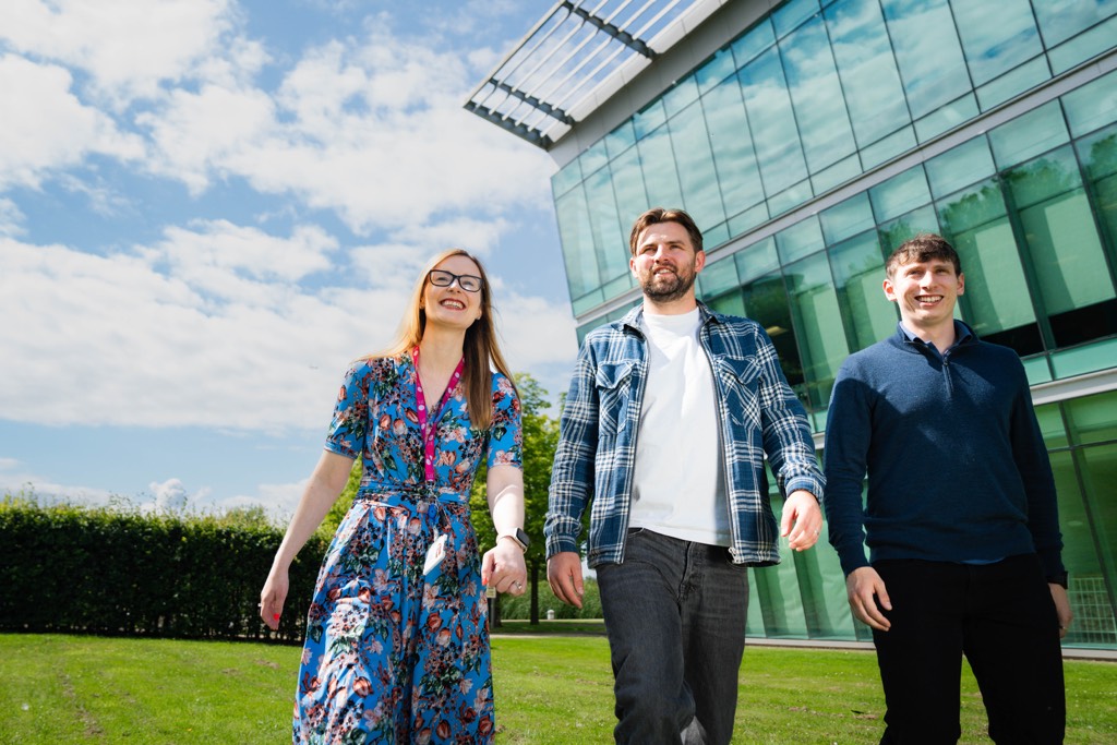 Three colleagues walk together across a grassy area outside Sky’s Livingston office, wearing smart-casual outfits including a patterned dress, a checked overshirt layered over a light top with dark jeans, and a knit jumper paired with dark trousers, with a modern glass-fronted building and blue sky behind them.