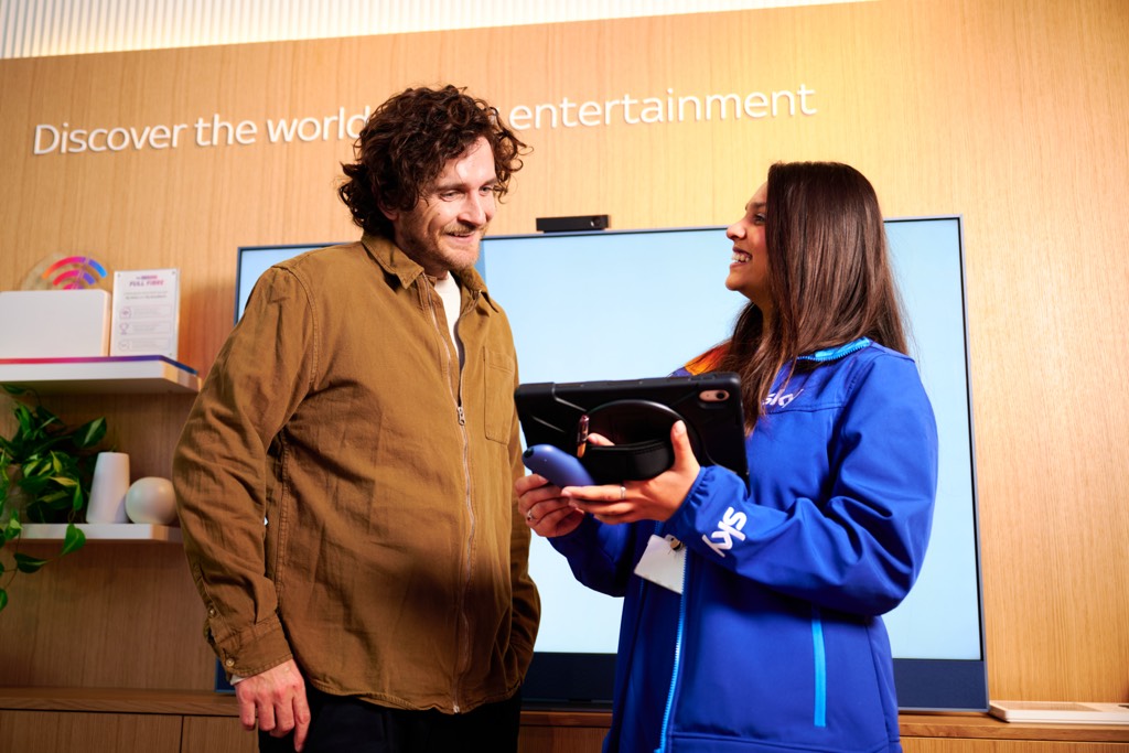 A customer stands on the left speaking with a Sky retail employee wearing a blue Sky‑branded jacket and lanyard, who is holding a handheld device. They are positioned in front of a large screen inside a Sky retail‑style environment, with shelving, product displays, plants, and wall text reading “Discover the world of entertainment” visible in the background.