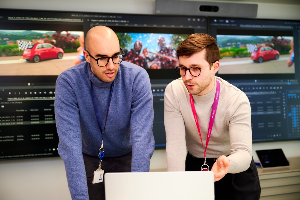 Two Sky colleagues collaborate at a workstation, leaning over a laptop while discussing work. Large monitors behind them display video editing software with multiple clips and timelines, suggesting a media or production environment inside a Sky office.