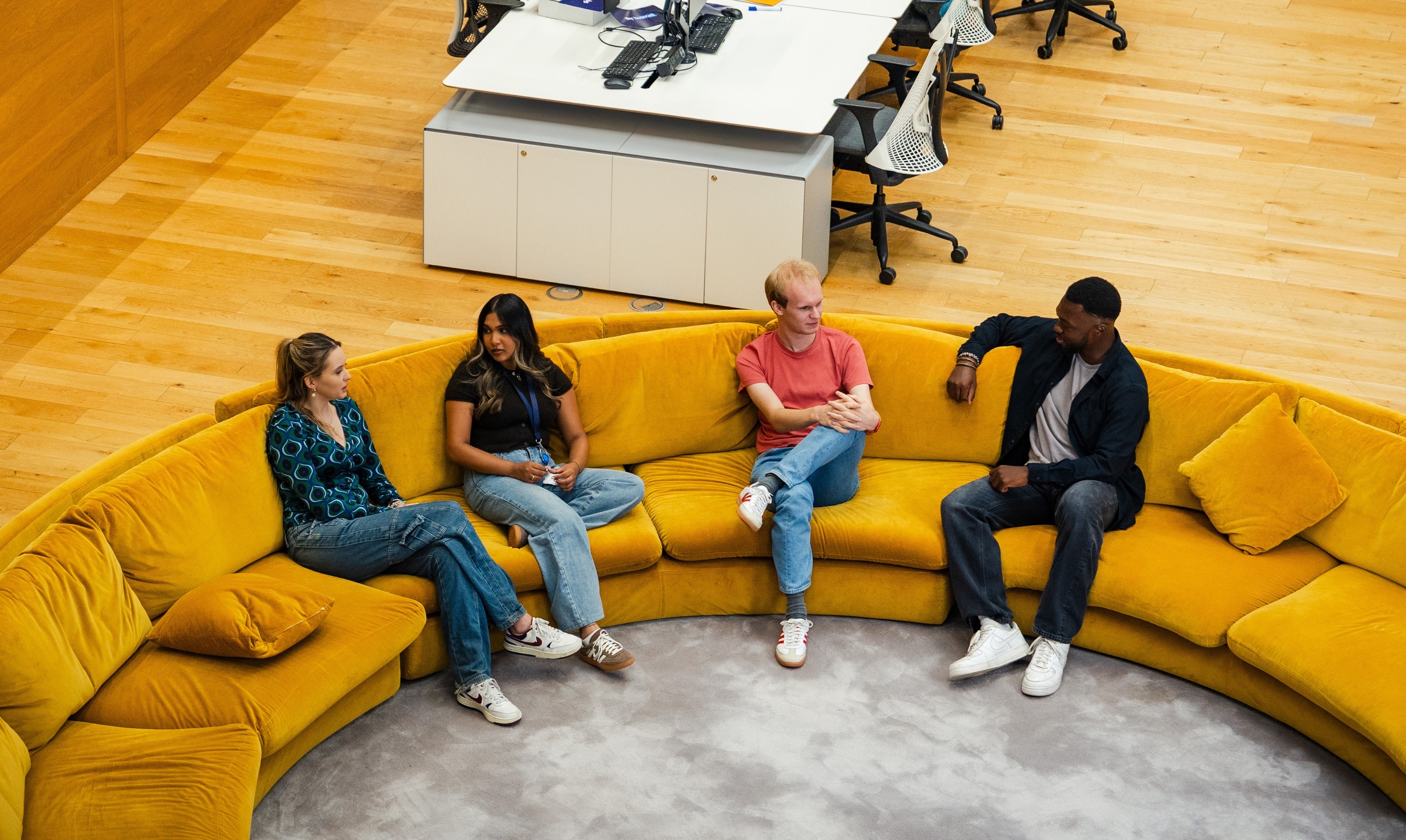 A group of colleagues sit and talk on a large curved yellow sofa in an open‑plan Sky office. The space features wooden flooring, modern desks with computer monitors in the background, and a collaborative lounge area designed for informal meetings and discussion.