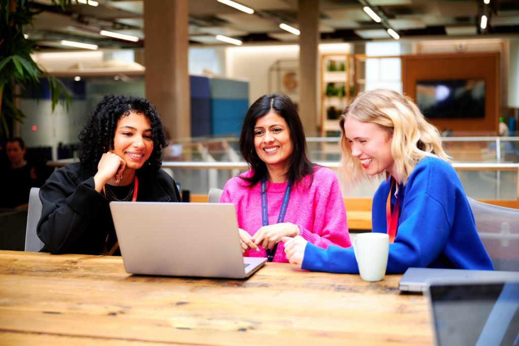 Three colleagues sit together at a large wooden table in an open‑plan Sky office, collaborating around a laptop. The workspace features exposed columns, modern lighting, indoor plants, and informal seating areas in the background, creating a bright and collaborative working environment.