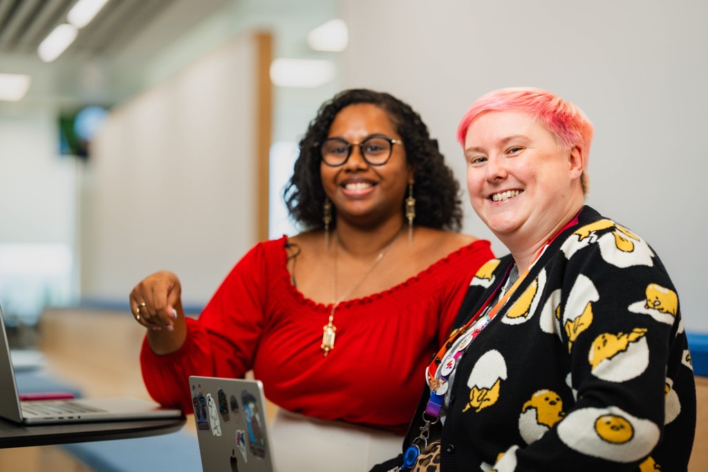 Two colleagues sit side by side at a table in a Sky office, working together on a laptop. One gestures toward the screen while the other looks on, with a bright corridor, glass panels, and modern office lighting visible in the background.