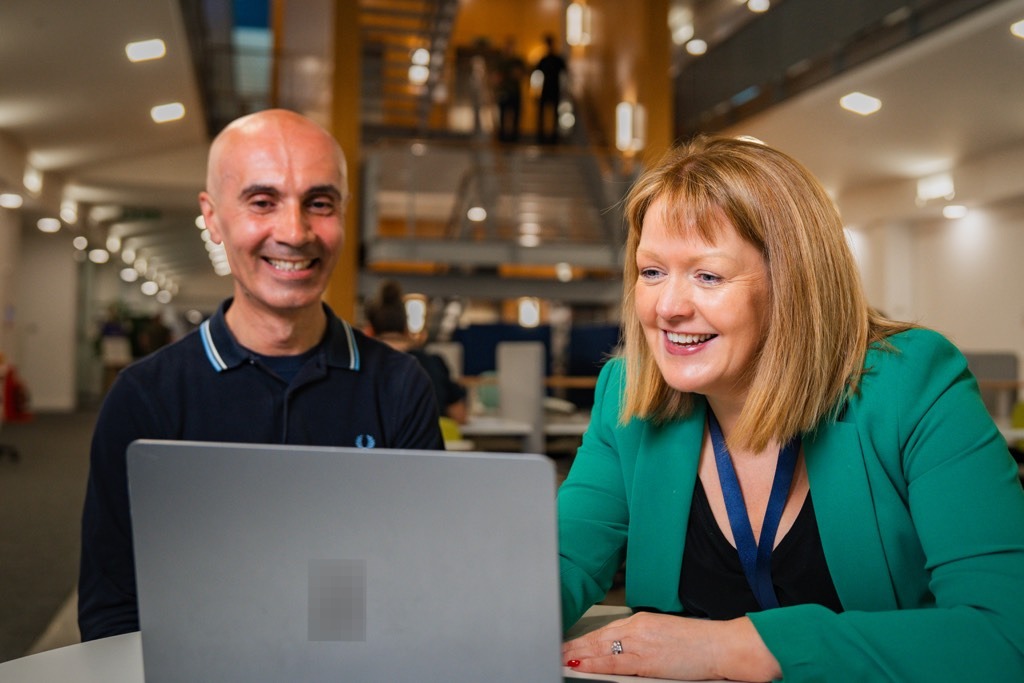 Two colleagues sit side by side at a table in a modern Sky office, working together on a laptop. The workspace features open seating, soft lighting, and a wide staircase with glass railings visible in the background, creating a collaborative and contemporary office environment.