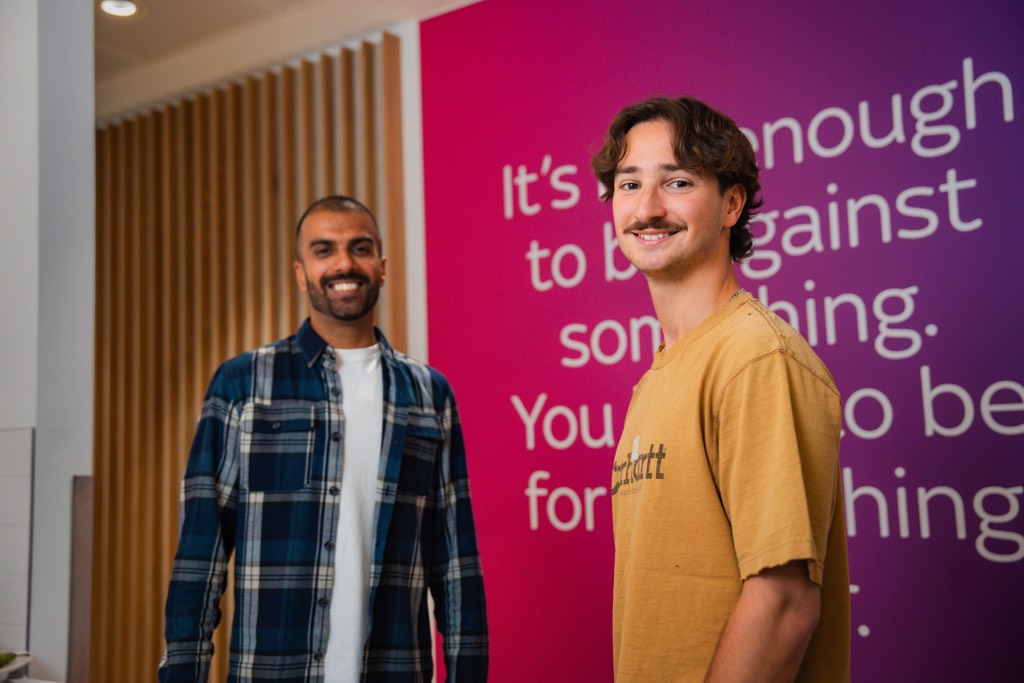 Two colleagues stand together in a modern Sky office space, positioned in front of a bold pink wall featuring large white text that reads, “It’s not enough to be against something. You have to be for something better.” One wears a checked shirt layered over a light top, while the other wears a casual short‑sleeve T‑shirt. Vertical wooden slats and contemporary interior finishes frame the setting, creating a clean and branded workplace environment.