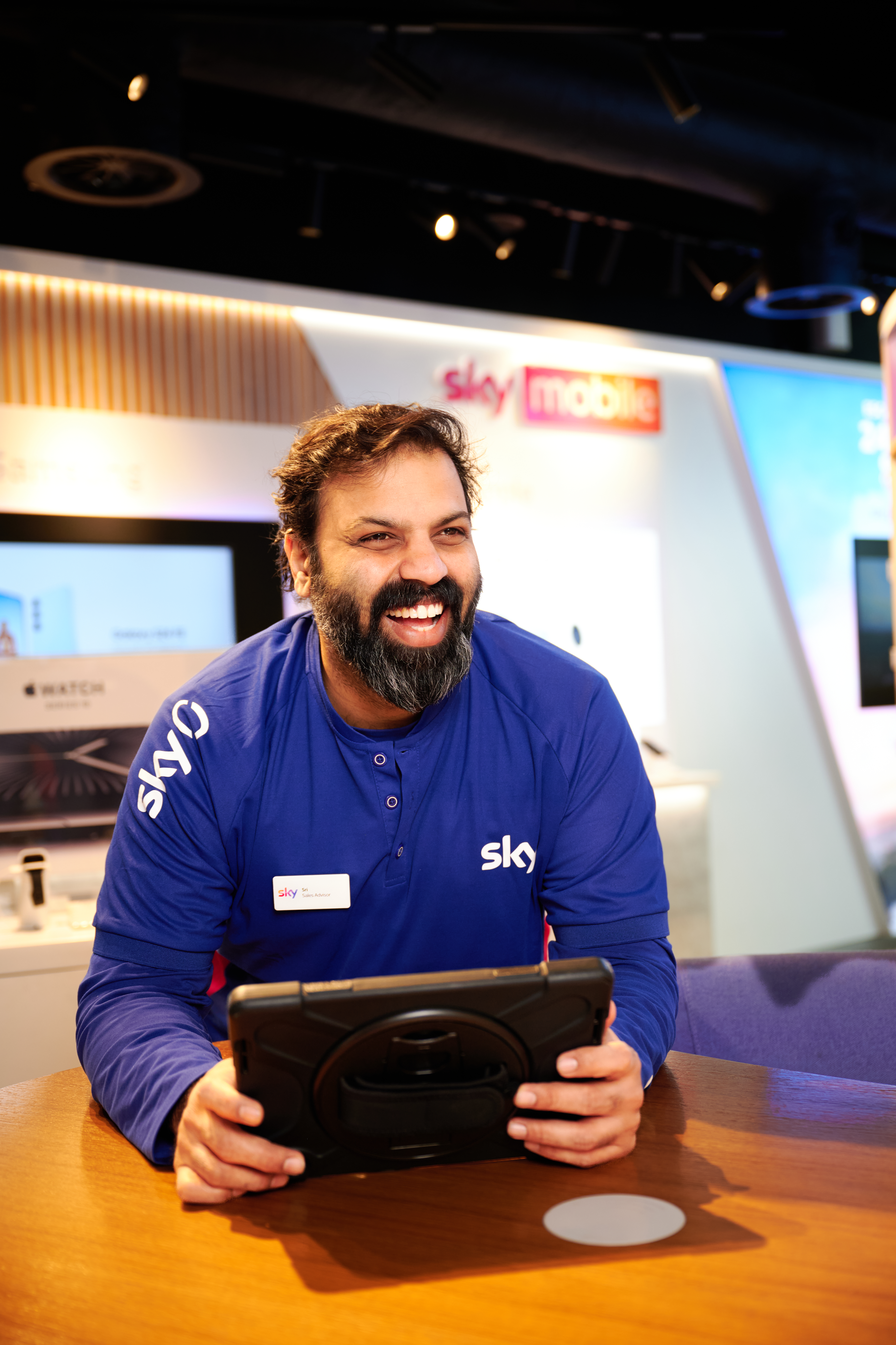 A Sky retail employee stands inside a Sky store, holding a tablet at a counter while reviewing information on the screen. The setting features branded displays, warm lighting, and Sky Mobile signage visible in the background, creating a modern customer‑facing retail environment.