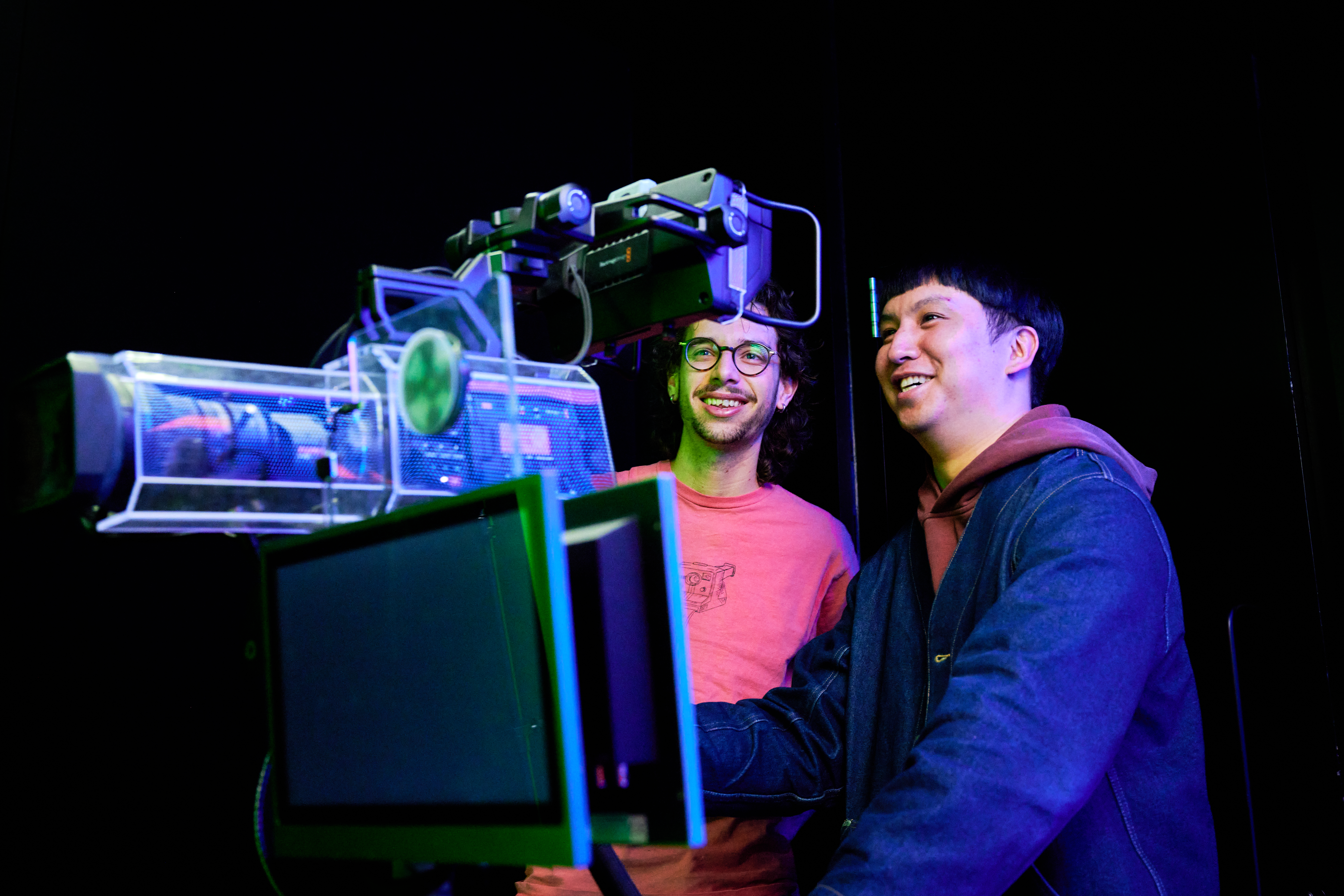 Two colleagues operate professional filming equipment in a dark studio environment, working together at a camera rig with mounted monitors and lighting. The scene is illuminated by coloured studio lights, highlighting the technical setup and creating a focused content‑production atmosphere.
