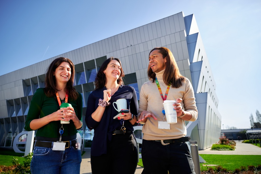 Three Sky colleagues walk together outside a modern Sky office building on a sunny day, holding takeaway drinks and chatting as they move along a landscaped path. The setting features a contemporary glass‑fronted building, green lawns, and clear blue sky, conveying a relaxed and sociable workplace environment.