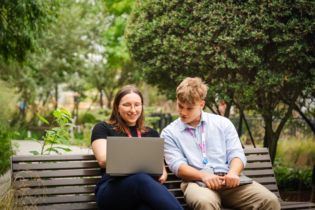Two Sky colleagues sit side by side on a wooden bench in a landscaped outdoor area, working together on a laptop. Trees, shrubs, and planted greenery surround the seating area, creating a calm, campus‑style setting that blends work and nature.