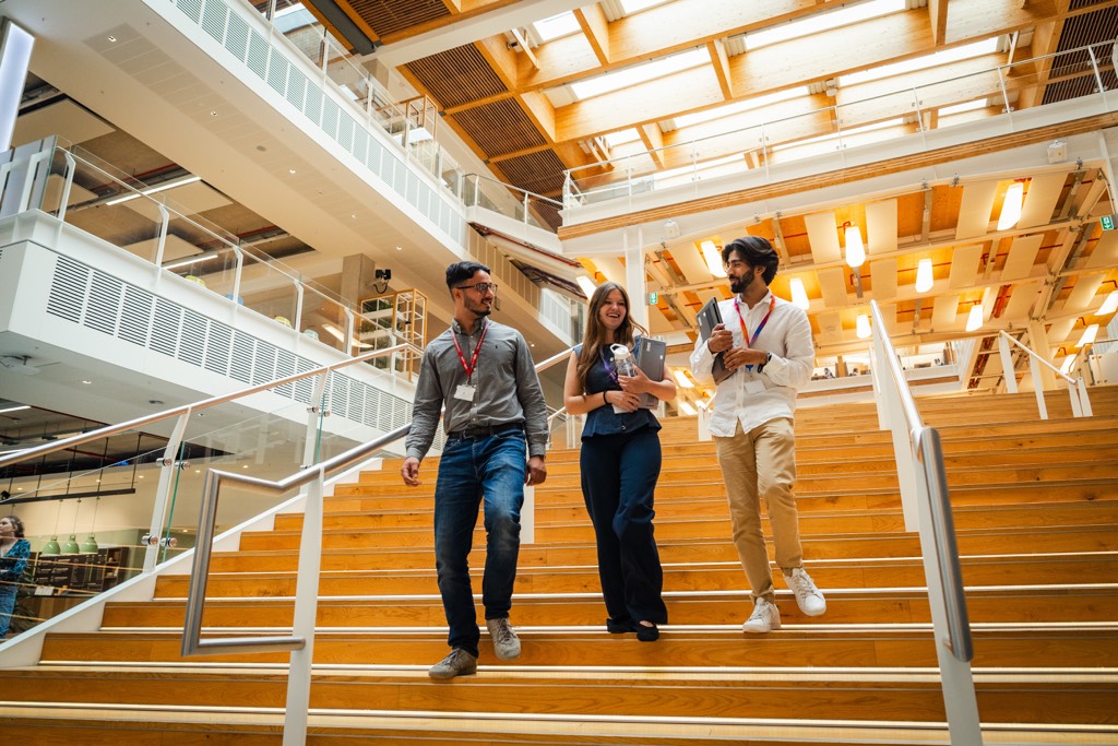 Three colleagues walk down a wide wooden staircase inside a bright, modern Sky office, talking together while holding notebooks and mobile devices. The open atrium features glass balustrades, exposed wood beams, warm overhead lighting, and multiple levels visible in the background, creating a spacious and collaborative workplace environment.