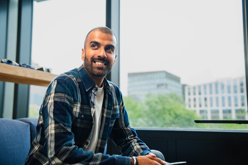 A colleague sits on a low sofa in a modern Sky office, holding a smartphone with both hands. They wear a checked overshirt layered over a light top and jeans. Large windows behind them let in natural daylight, with structural beams and greenery visible outside, creating a relaxed and contemporary workplace setting.
