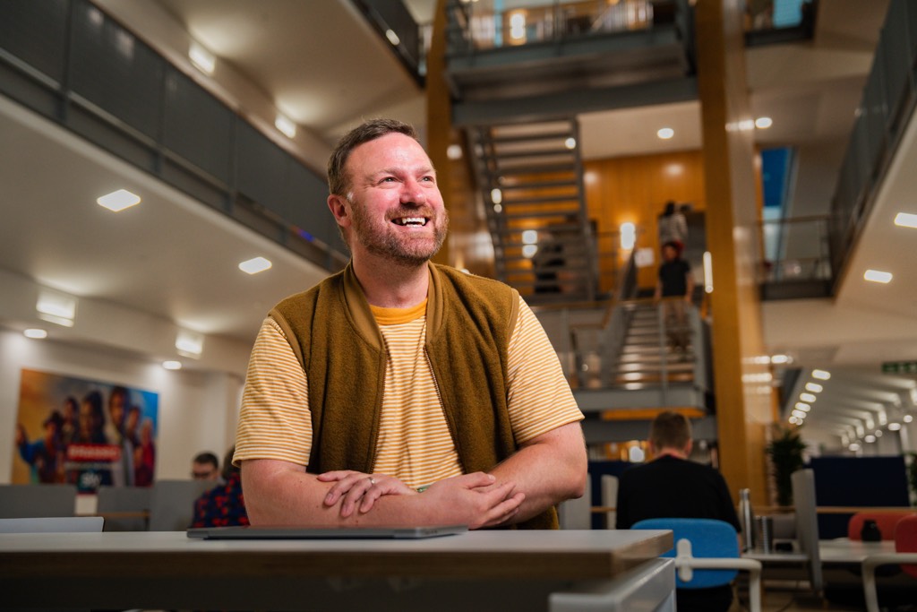 A colleague stands at a high table inside a bright, open‑plan Sky office, resting their arms on the surface while wearing a striped T‑shirt layered with a sleeveless vest. The background shows a spacious atrium with wooden staircases, glass railings, multiple floors, and other colleagues working and moving through the space, creating a busy and collaborative workplace environment.