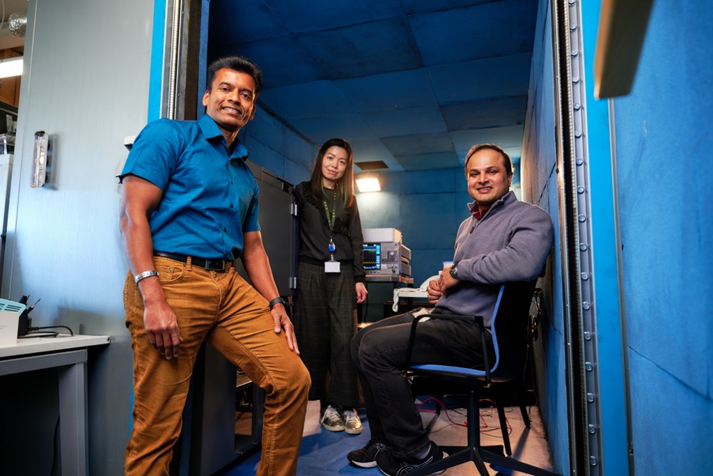 Three colleagues are positioned inside a compact, blue‑lined technical room, surrounded by specialist equipment and control panels. One stands near the doorway, another stands beside monitoring devices, and a third sits on a swivel chair, suggesting a controlled testing or recording environment within a Sky facility.