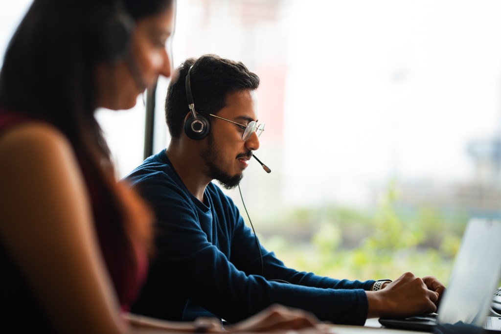 Two colleagues work side by side at a desk in a Sky office, focused on laptops. One wears an over‑ear headset while typing, and the other sits slightly in the foreground. Large windows allow natural daylight into the space, with greenery visible outside, creating a calm and modern working environment.