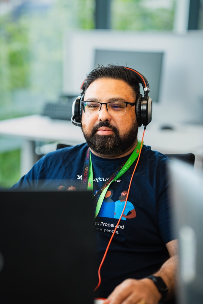 A colleague sits at a desk in a Sky office, wearing over‑ear headphones and working on a laptop. A green lanyard is visible, and the background shows computer monitors, large windows, and greenery outside, creating a calm, modern technology‑focused workspace.