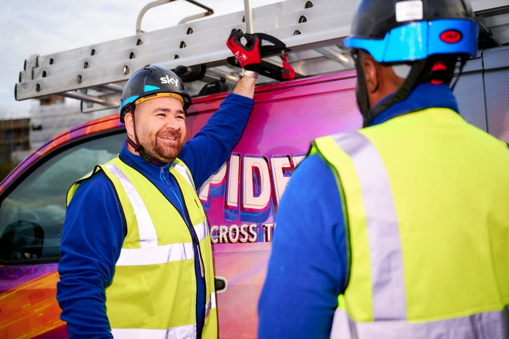 Two Sky field engineers stand beside a branded Sky service van, wearing high‑visibility vests, blue helmets, and protective gloves. One reaches up to adjust a ladder secured to the roof rack while the other stands nearby, with tools and safety equipment visible, suggesting preparation for on‑site installation or maintenance work.