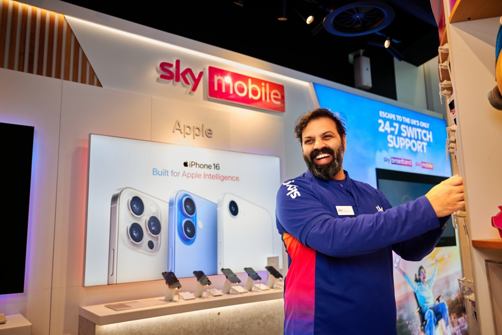 A Sky Mobile retail employee stands inside a Sky store, adjusting a display fixture near a product wall. Behind them, illuminated signage reads “sky mobile” and “Apple,” with a display for iPhone 16 stating “Built for Apple Intelligence.” Smartphones are arranged on a counter below the screen, and additional digital signage advertises “24/7 Switch Support,” creating a modern, branded retail environment.