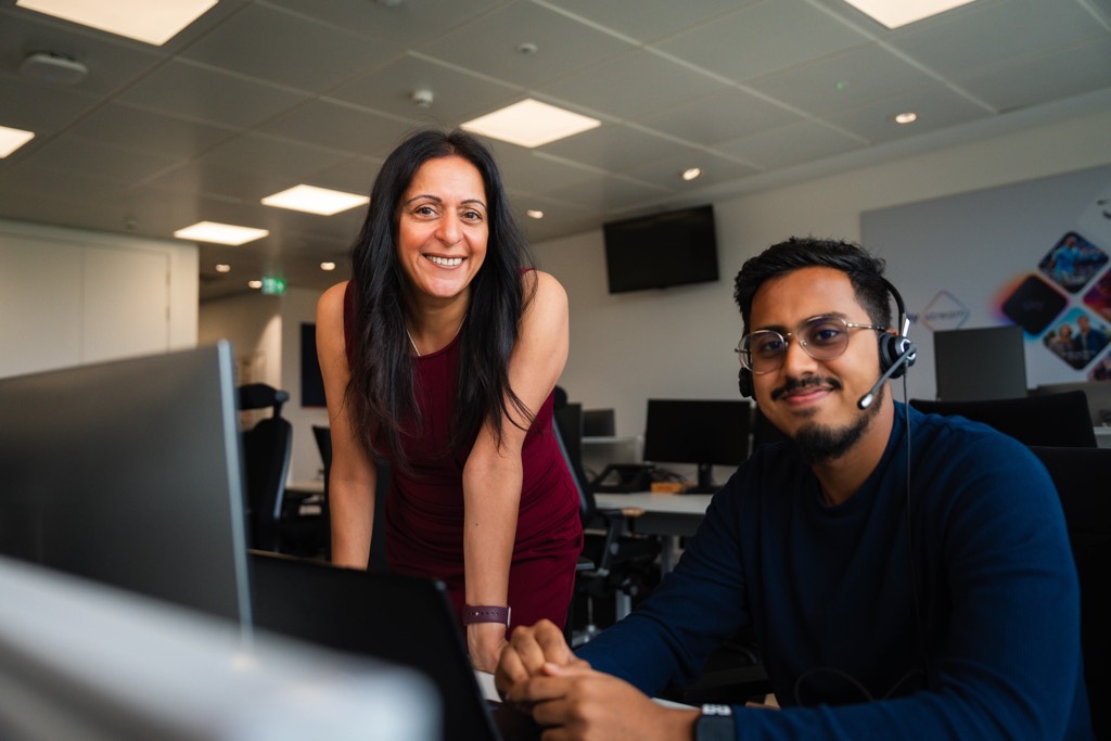 Two colleagues collaborate at a workstation inside an open-plan Sky office. One stands leaning forward beside a desk while the other sits at a computer wearing a headset, both focused on the screen. The space features rows of desks, monitors, overhead lighting, and wall-mounted screens, creating a modern, professional working environment.