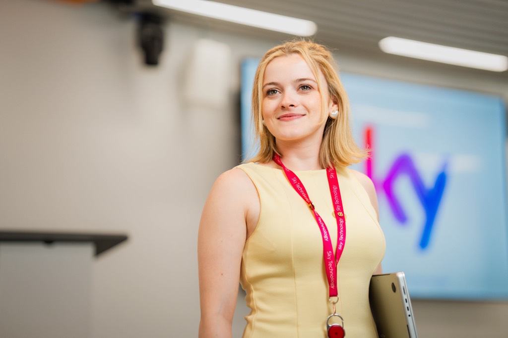 A colleague stands in a modern Sky office or meeting space, wearing a sleeveless yellow dress and a Sky-branded lanyard, holding a laptop at their side. Behind them, a large screen displays Sky branding, with ceiling lighting and presentation equipment visible, suggesting a professional presentation or collaborative work setting.