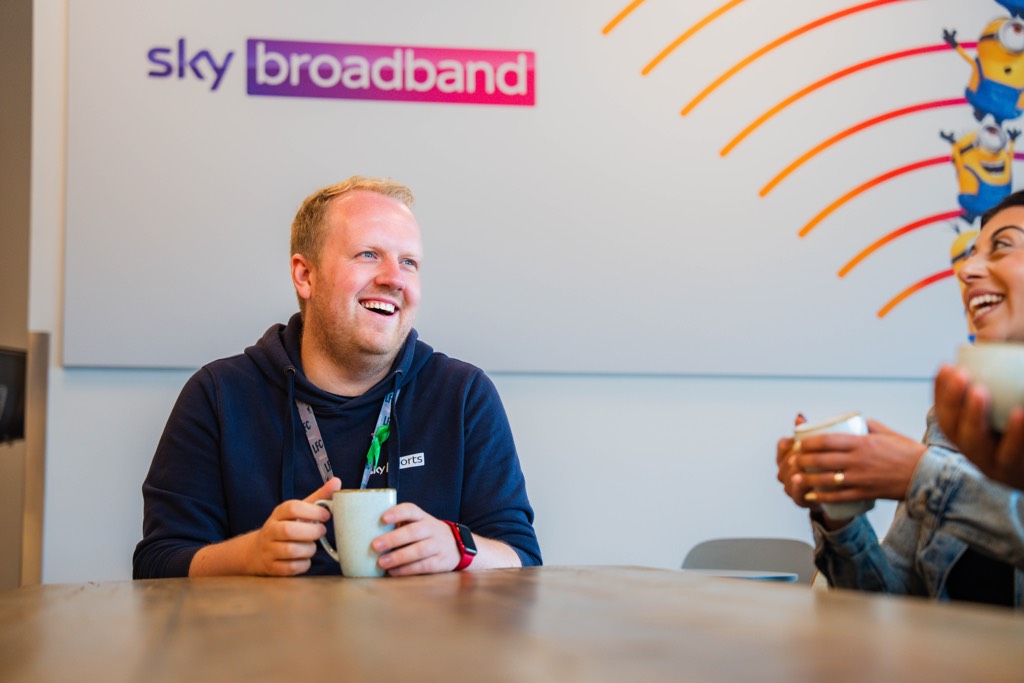 Two colleagues sit at a meeting table inside a Sky office, each holding a mug during an informal conversation. Sky Broadband branding is visible on the wall behind them, along with playful graphic elements, creating a relaxed, collaborative workplace setting.